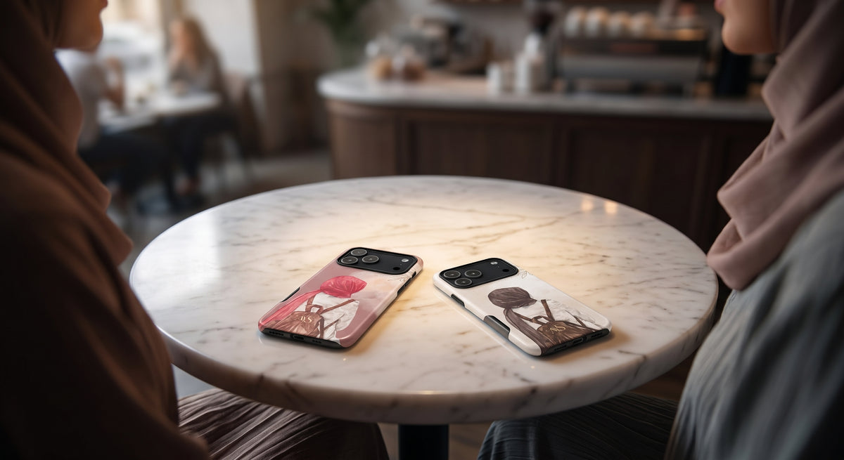 Rose Serenity and Mocha Rose hijabi tough phone cases with illustrated designs resting on a round white marble cafe table, with two women in hijabs seated on either side in a softly blurred coffee shop background.
